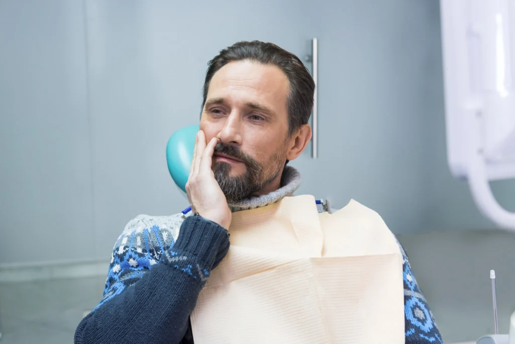 Adult man holding his jaw at the dentist, showing post-surgery discomfort after wisdom tooth removal